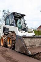 A skid steer loader with a front bucket sits on muddy ground. Leafless trees and a red roofed building partially hidden by shrubs are visible. photo