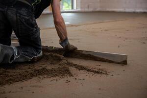 A worker kneels on a construction site, using a straightedge tool to level sand or concrete. Unfinished walls and an open window provide natural light. photo