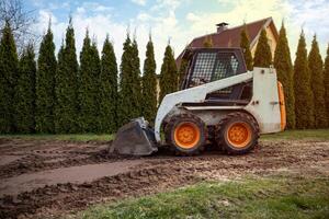 A skid steer loader with orange wheels rests on a dirt path with visible tire tracks. Tall evergreen trees and a house with a sloped roof are nearby. photo
