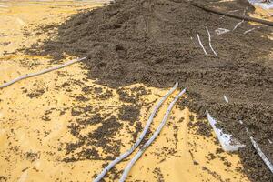 A construction site showing a yellow plastic sheet partially covered by gravel or concrete mix, with white pipes scattered across the surface. photo