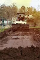 A man uses a small bulldozer to level a dirt path. Trees and fencing border the area, with freshly turned soil in the foreground and a structure visible. photo