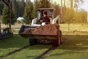 A man drives a skid steer loader carrying soil in an open grassy area. Tracks are visible on the grass, with a fence, forest, and small structure nearby. photo