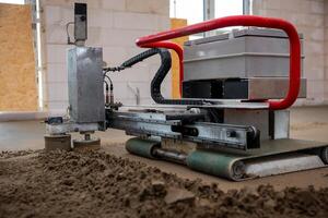 A specialized machine with a red handle levels sand on a concrete floor. Surrounding walls are unfinished, with concrete blocks and plywood openings. photo