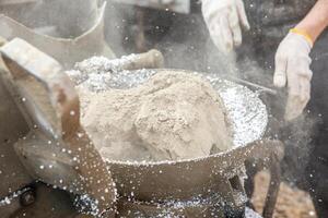 Powdered material and white granules mix in a metal container, with gloved hands nearby. Dust particles are visible, indicating an industrial process. photo