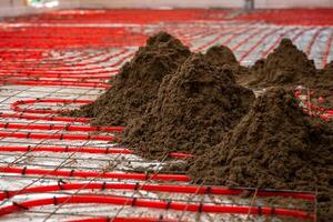 Red underfloor heating pipes in a grid pattern secured with metal mesh, with piles of Estrich concrete ready for spreading and leveling. photo