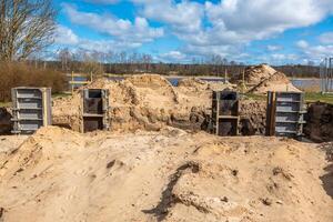 Construction site featuring metal framework in sandy soil, piles of sand, and a body of water bordered by sparse trees under a partly cloudy sky. photo