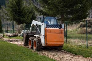 An orange and white skid steer loader rests on a dirt path with its bucket down. The area features tire tracks, disturbed soil, and tall evergreen trees. photo