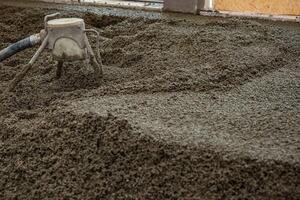 Freshly poured concrete floor in an indoor construction site, with a leveling tool connected to a hose in use. Partially visible wall and window frame. photo