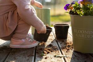 A child in pink shoes and light clothing works with soil in black pots on a wooden deck. A green container with purple and yellow flowers is nearby. photo
