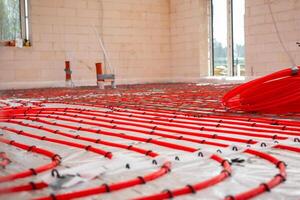 Interior of a construction site showing red underfloor heating pipes in a grid pattern over reflective insulation, with light brick walls and window openings. photo