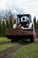 A man in a black cap and shirt operates a skid steer loader carrying soil across grass. Tracks are visible, with conifer and deciduous trees in the background. photo