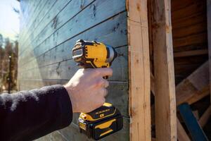 A person uses a yellow and black cordless drill to secure dark wooden planks. The scene includes wooden elements and natural light in an outdoor setting. photo