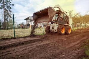 A skid steer loader with orange wheels spreads soil on a dirt path near a wire fence, with residential buildings and sparse trees in the distance. photo
