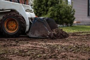 A skid steer loader with orange wheel rims moves soil with its bucket. Nearby are a green bush, a building with horizontal siding, and excavated ground. photo