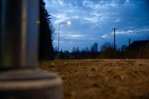 A dirt pathway lit by streetlights under a deep blue evening sky, with power lines, a utility pole, tree silhouettes, and a blurred metallic object in view. photo