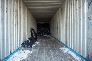 White walled shipping container with ribbed structure, wooden floor, black plastic bags at the far end, and scattered white particles on the floor. photo