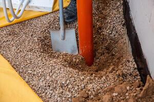 A construction site showing a partially buried orange pipe in gravel, an upright metal shovel, a worker's boot, and a yellow protective sheet. photo