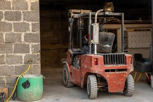 A red forklift sits partially inside a building with unfinished brick walls and a concrete floor. A green barrel with a yellow hose is near the entrance. photo