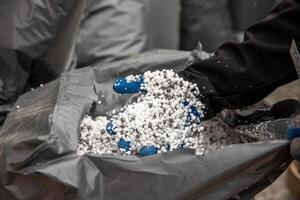 A close up of a gloved hand holding small white granules, likely fertilizer, scooped from a large gray bag in an industrial or agricultural setting. photo