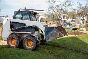A skid steer loader with orange wheels carries soil in its raised bucket on a grassy surface. Nearby are houses, a swing set, and leafless trees. photo