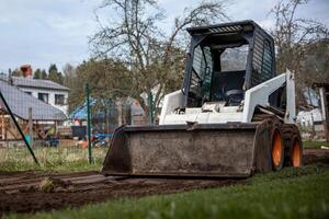 A skid steer loader with orange wheels and a white body is parked on a dirt path. Nearby are trees, a fenced area, and houses with slanted roofs. photo
