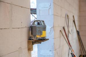 A makeshift device wrapped in duct tape sits on a wooden shelf against a light brick wall, surrounded by tools and an unfinished workshop environment. photo