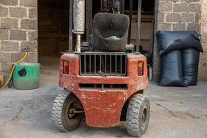 A red forklift with visible wear stands near a stone walled structure with an open doorway. A green container and black bags are placed nearby on concrete ground. photo
