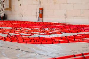 Red underfloor heating pipes arranged on reflective insulation, secured with black fasteners. Partially finished wall with wiring and vertical orange pipe visible. photo