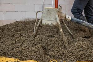 A concrete screed tool with a tripod structure and hose rests on lightweight aggregate concrete. A worker stands nearby near a white brick wall and red pipe. photo