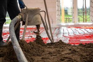 Red heating pipes are arranged on the floor as a worker pours screed concrete using a pump inside a building under construction with open windows. photo