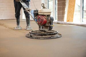 A worker operates a power trowel with a red engine to smooth concrete flooring. Concrete block walls and large windows are visible in the construction site. photo