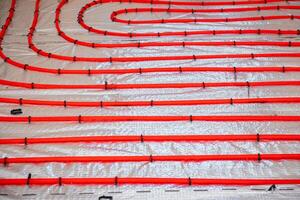 Bright red underfloor heating pipes arranged in loops, secured with black fasteners on a silver insulation layer during the installation phase. photo