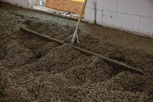 An indoor construction site shows wet concrete being leveled with a rake like tool. Unfinished white block walls and a wooden panel are visible. photo