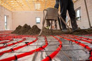 A construction worker installs red underfloor heating pipes in a systematic layout and pours Estrich concrete in a building with unfinished walls and roof frame. photo