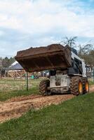 A skid steer loader with a raised bucket of soil sits on a muddy dirt path, bordered by a wire fence and a wooden structure under an overcast sky. photo