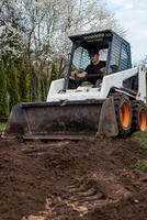 A man uses a skid steer loader with orange wheels and a front bucket to move soil. Flowering trees with white blossoms and hedges are visible behind. photo