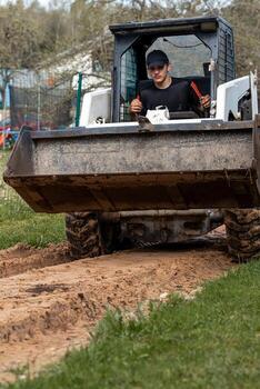 A man in a dark cap and shirt operates a skid steer loader with a bucket attachment on a dirt path. Grass, a wire fence, and structures are visible. photo