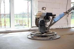 A power trowel machine smooths fresh concrete flooring in a building under construction with white brick walls, glass windows, and scaffolding visible. photo