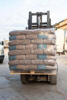 A forklift carries a pallet of clear wrapped bags on paved ground near a white van and brick building, indicating active material handling. photo