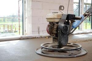 A power trowel machine smooths fresh concrete flooring inside a white brick structure with large windows and visible scaffolding outside. photo