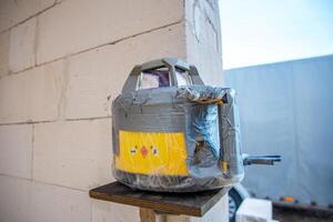 A yellow and gray surveying device wrapped in plastic and duct tape is mounted on a wooden platform near a light brick wall. A white vehicle and gray structure are visible. photo