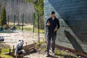 A man in dark clothing stands by a green wooden structure holding a smartphone and bottle. A black and white dog walks near a fenced dirt path with sparse trees. photo