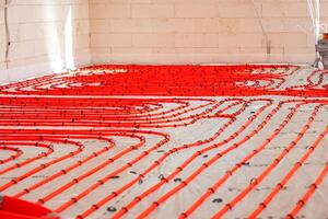 Interior of a construction site showing red underfloor heating pipes arranged in loops on a base layer, with light brick walls and visible electrical conduits. photo