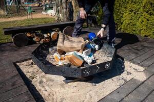 A hexagonal fire pit with decorative cutouts on a sandy base, surrounded by wooden decking, firewood on a log bench, and a person tending the fire. photo