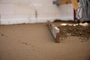 A screed tool levels freshly laid Estrich concrete indoors, with white walls, electrical outlets, and cables visible in the background. photo