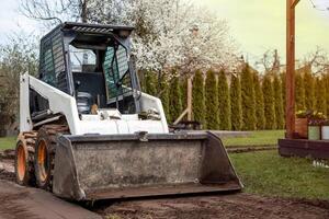 A skid steer loader with a front bucket sits on a dirt path, surrounded by grass, blooming white trees, tall hedges, and a wooden post with flower pots. photo