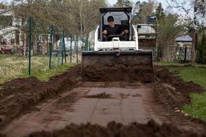 A man uses a white bulldozer to level soil on a narrow pathway bordered by grass, with a wire fence, houses, and trees visible in the distance. photo