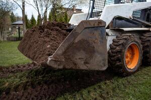 A white skid steer loader with orange wheels and a soil filled bucket on grass. Tire tracks, trees, a gazebo, and a building are visible nearby. photo