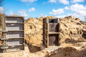 A construction site featuring two metal and wooden formwork structures on sandy ground, with mounds of sand and a wire fence under clear daylight. photo