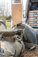 A construction site featuring a worn cement mixer, a van loaded with cement bags, gravel covered ground, sparse trees, and a distant fence. photo